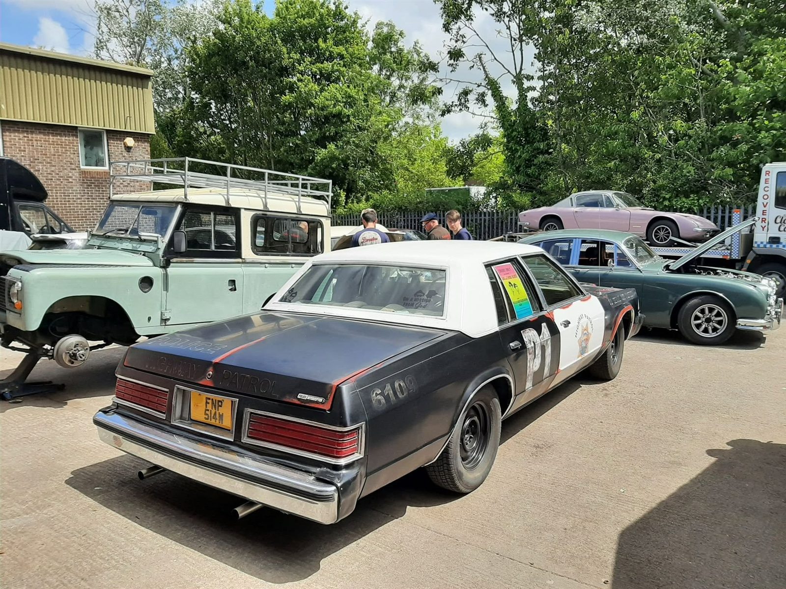 American saloon in highway patrol livery, with a Land Rover and other classics in a yard